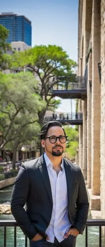 Modern architectural designer, male, 30s, bespectacled, short hair, casual wear, standing, San Antonio, Texas, River Walk, cypress trees, limestone buildings, iron balconies, vibrant street art, sunny